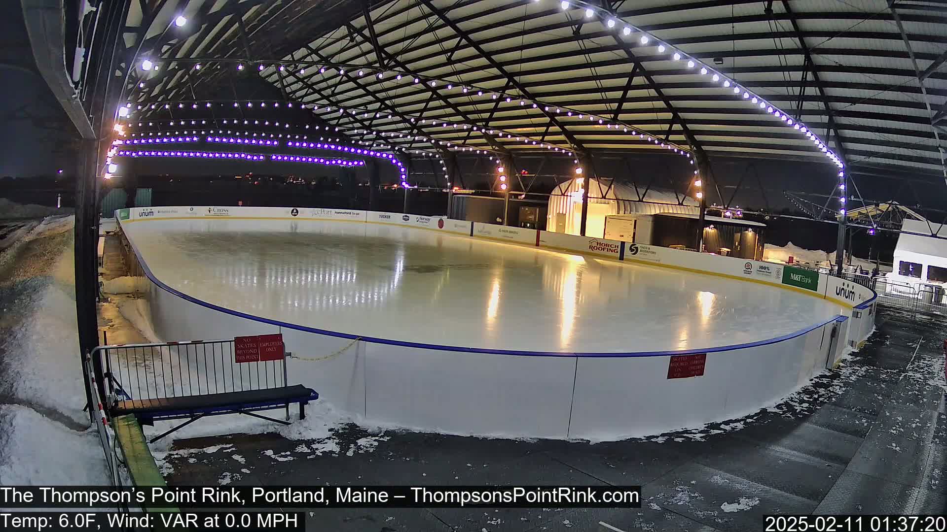 An outdoor ice rink at night, illuminated by lights strung across its covered structure, with snow on the ground around the rink.