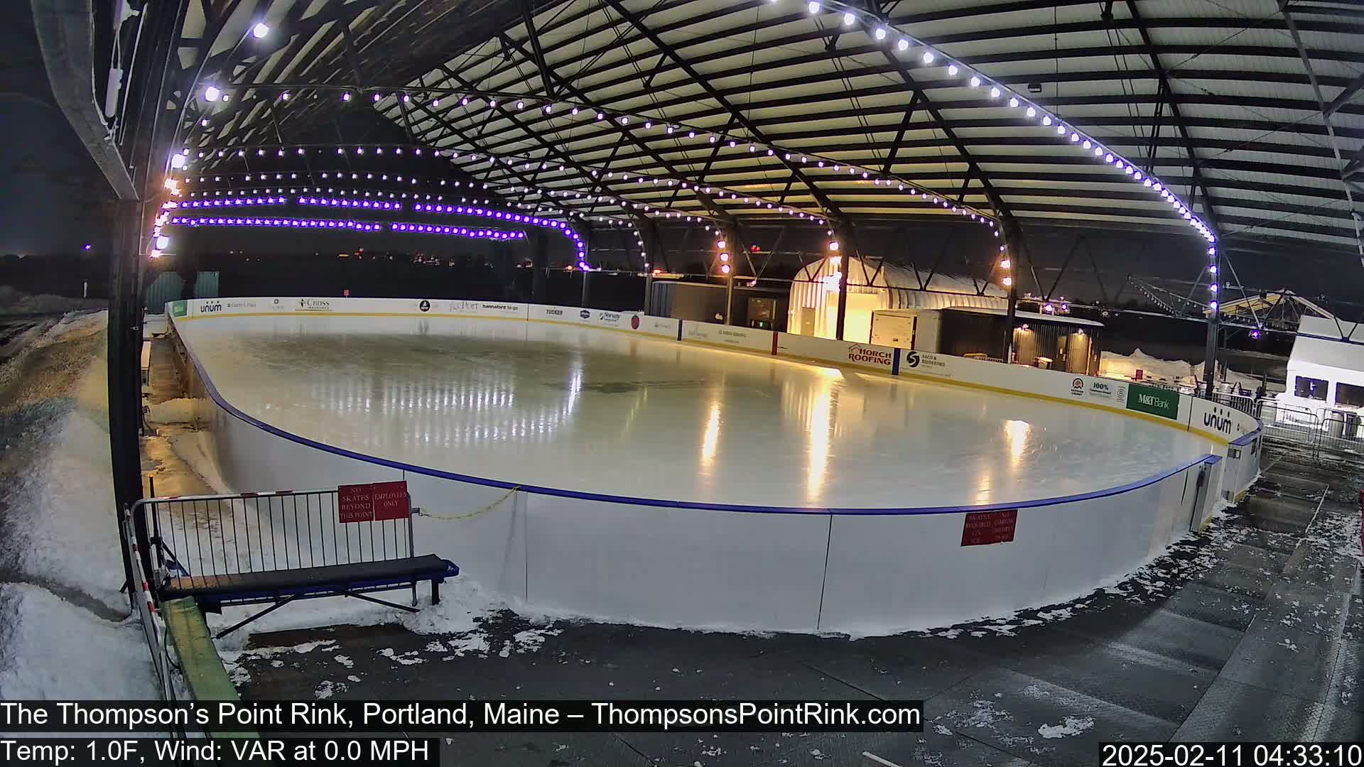 An outdoor ice rink, illuminated at night under a metal structure with string lights, is partially covered in snow.
