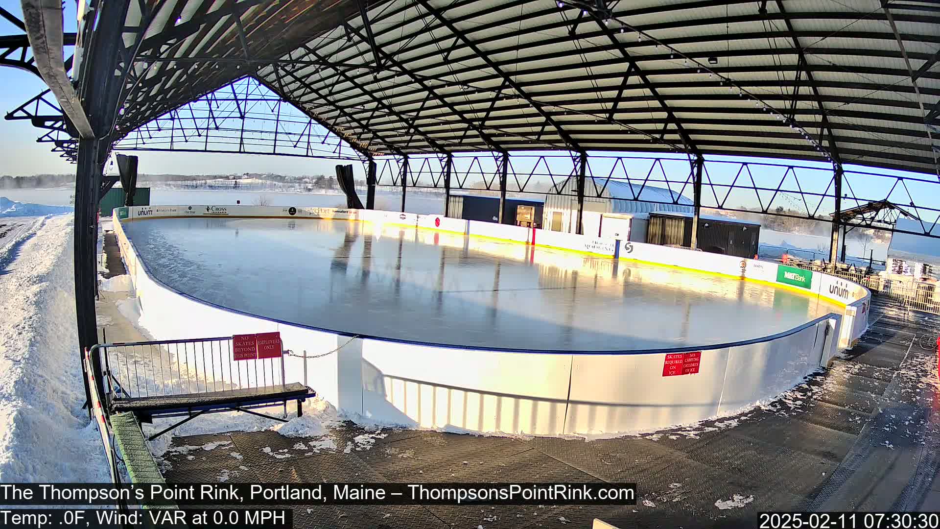 A sunny, snow-covered outdoor ice rink sits under a metal roof structure.