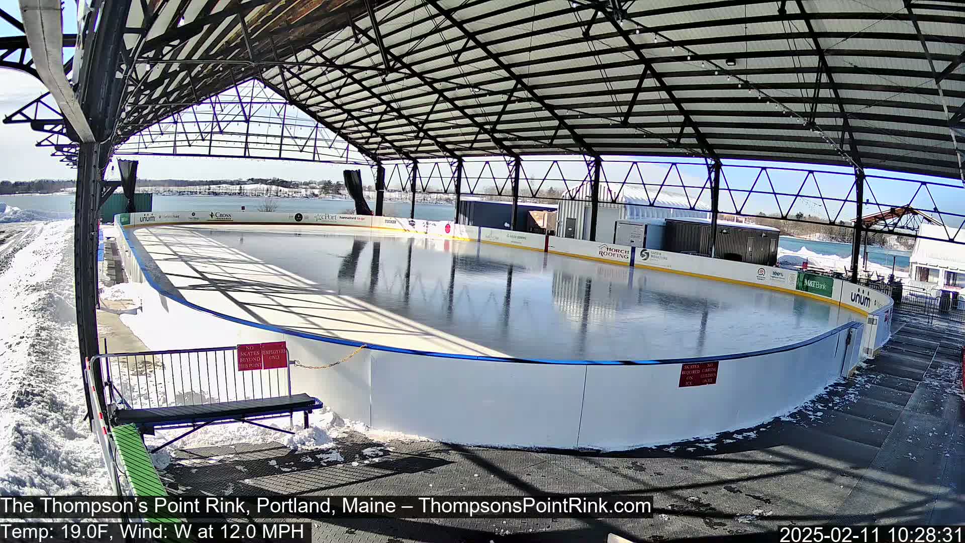 A sunny outdoor ice rink, partially covered by a metal structure, is surrounded by snow.
