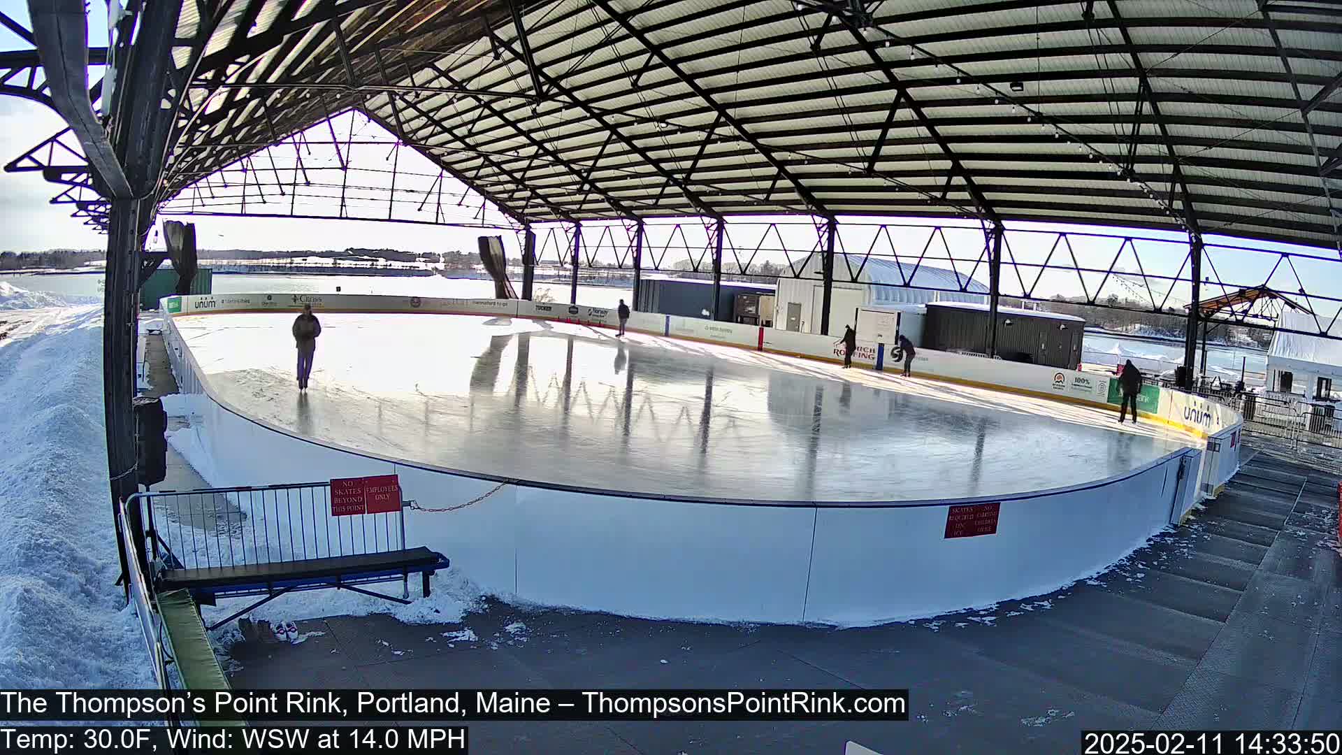 A sunny outdoor ice rink, partially covered by a metal structure, with several people ice skating on a clear, reflective surface; snow is visible on the ground around the rink.