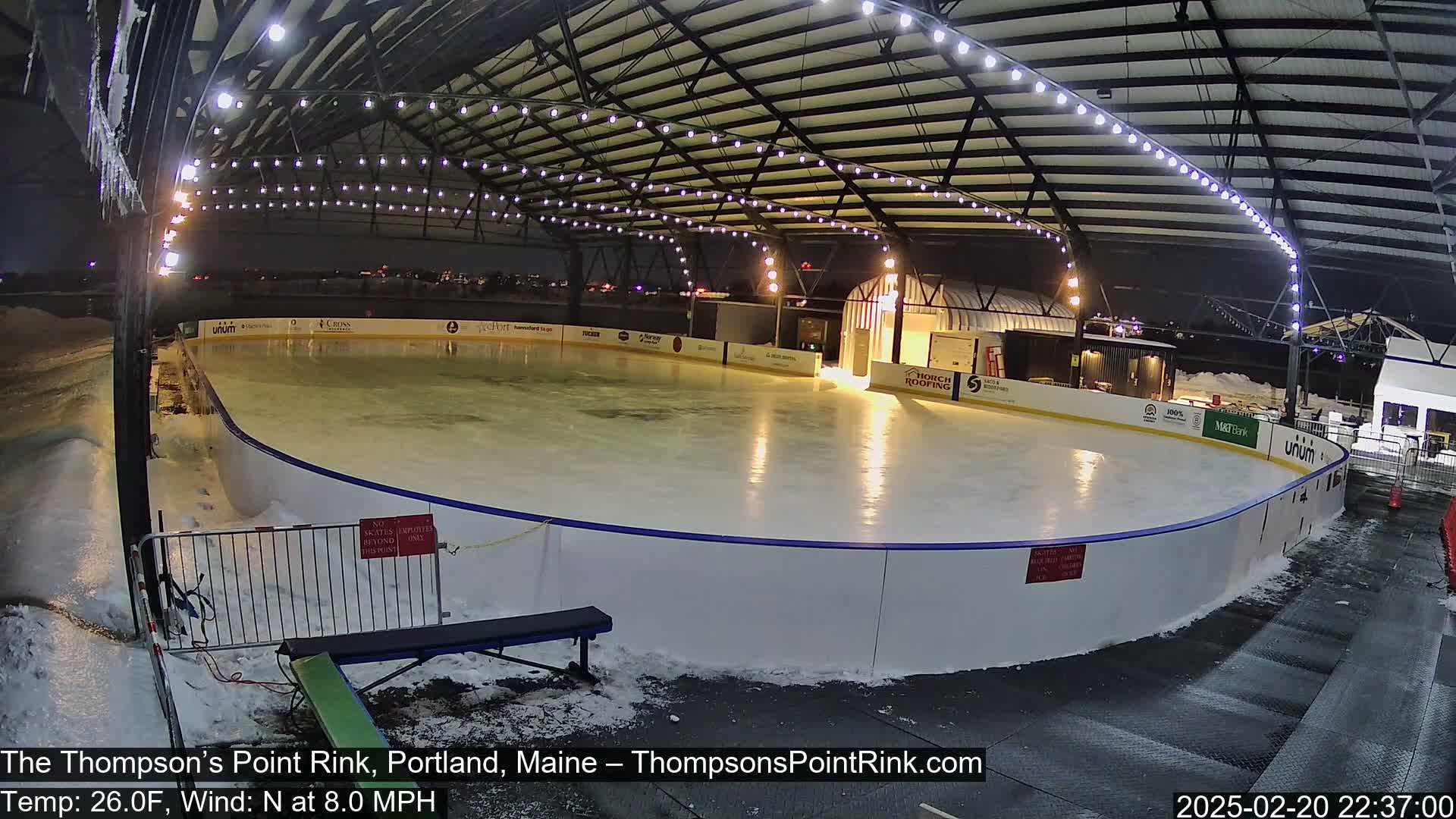 An outdoor ice rink is illuminated at night under a metal structure, with snow on the ground and an eight-mile-per-hour north wind at 26 degrees Fahrenheit.