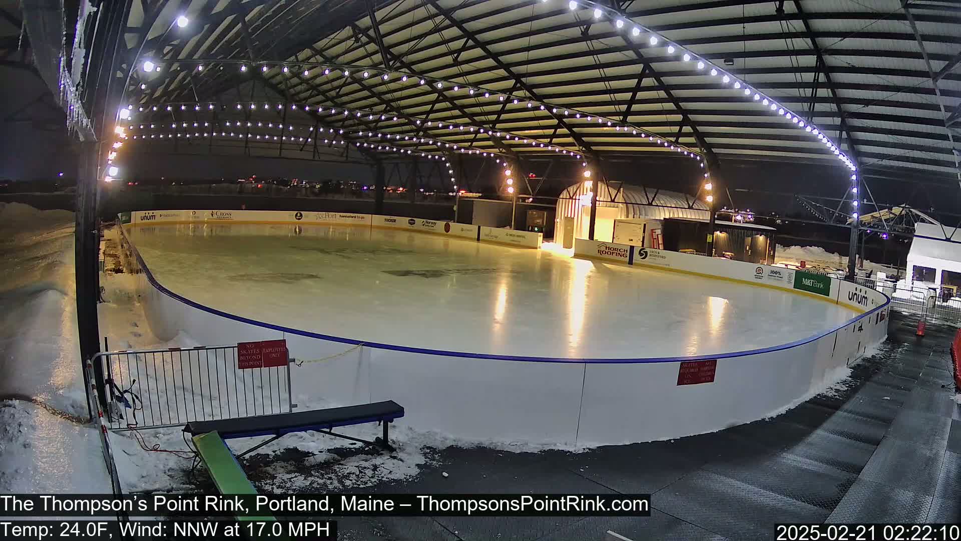 An outdoor ice rink, illuminated at night under a covered structure, is surrounded by snow in windy, 24-degree Fahrenheit weather.
