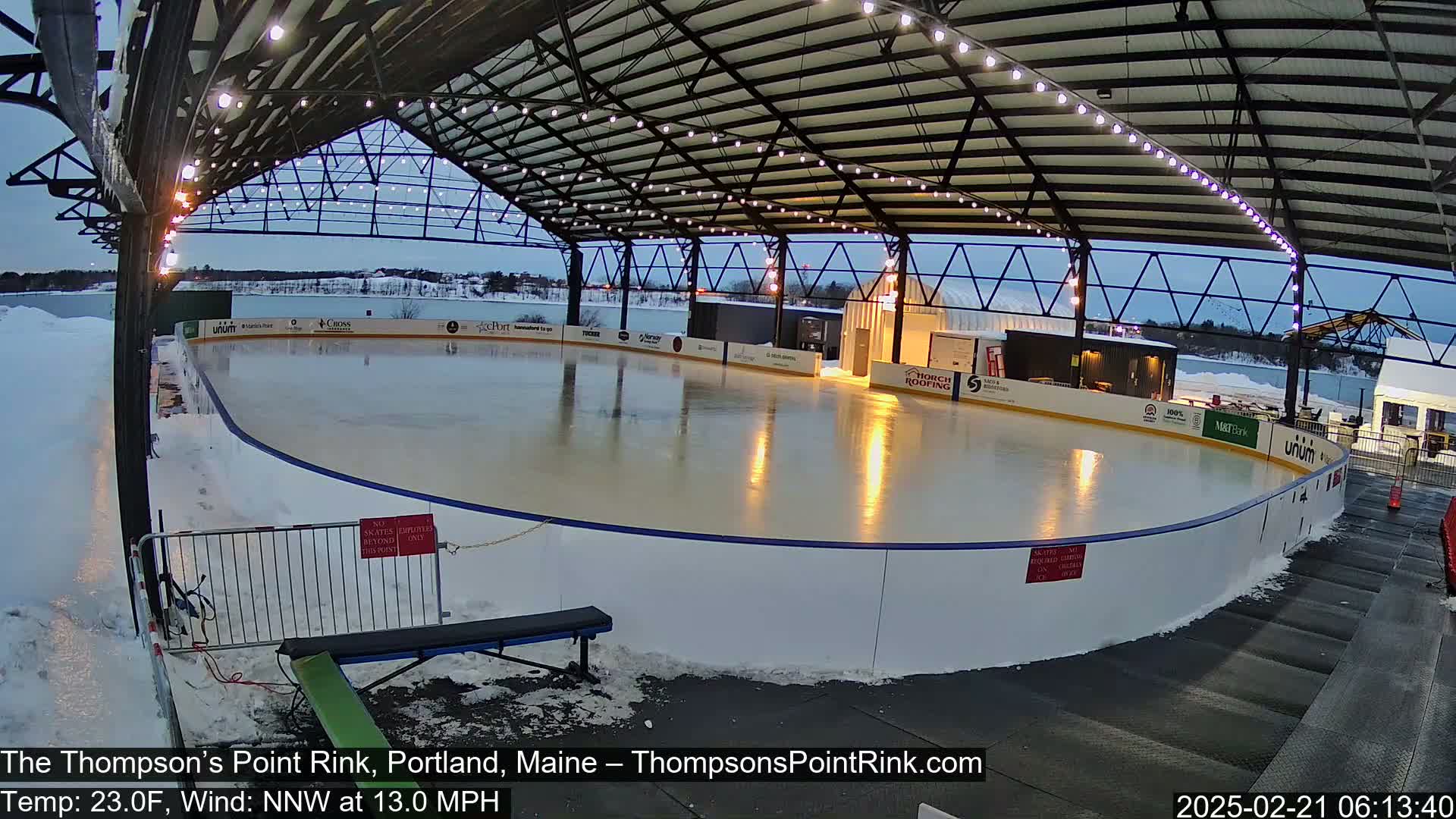 An outdoor ice rink, under a metal structure with string lights, is mostly clear of people, surrounded by snow and a bench.
