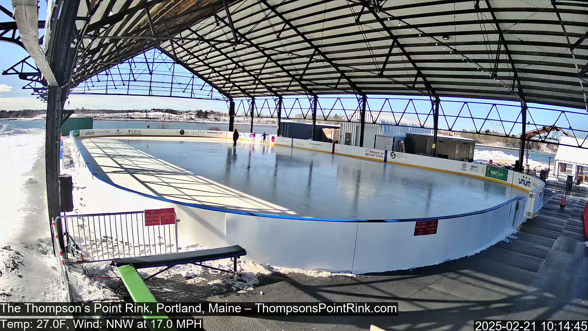 A partially covered outdoor ice rink with a few people ice skating under a sunny sky.