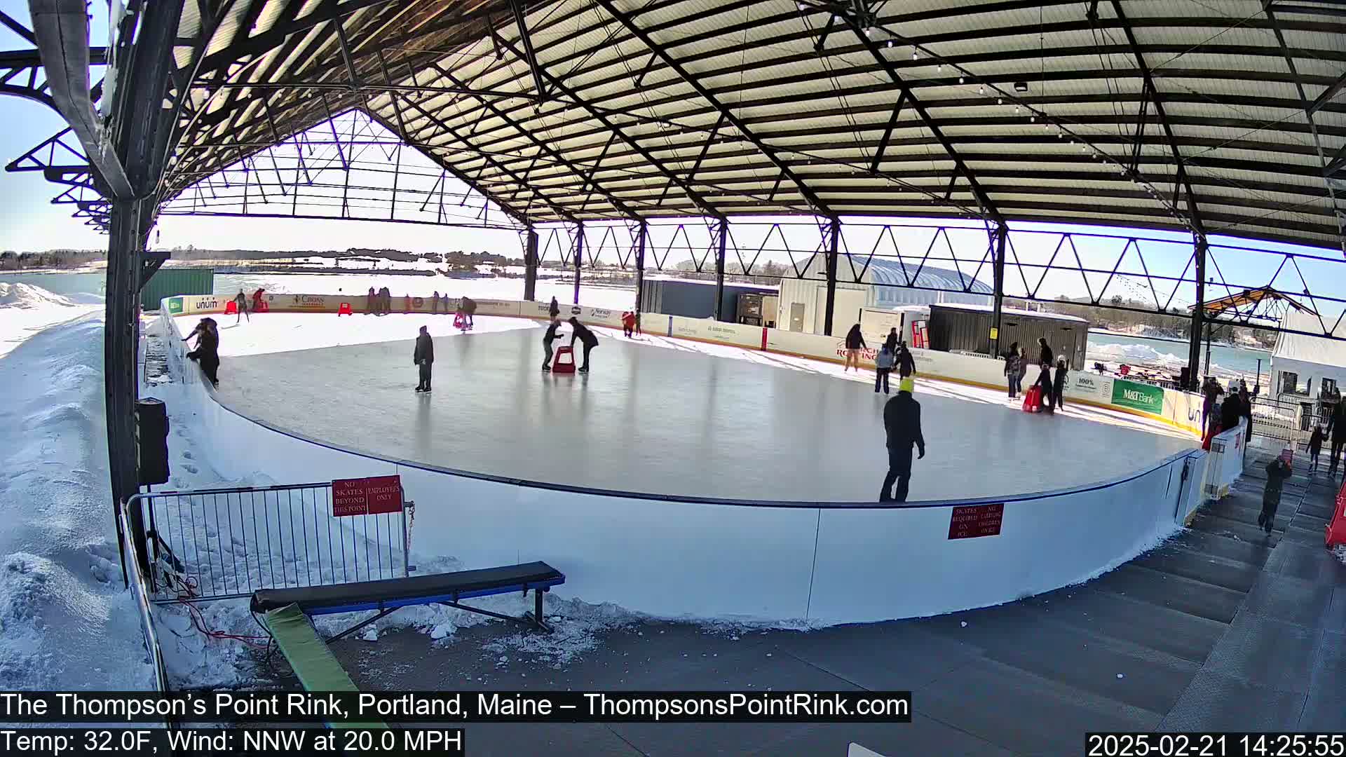 An outdoor ice rink, under a metal structure, is surrounded by snow on a sunny day with a 20 mph north-northwest wind.