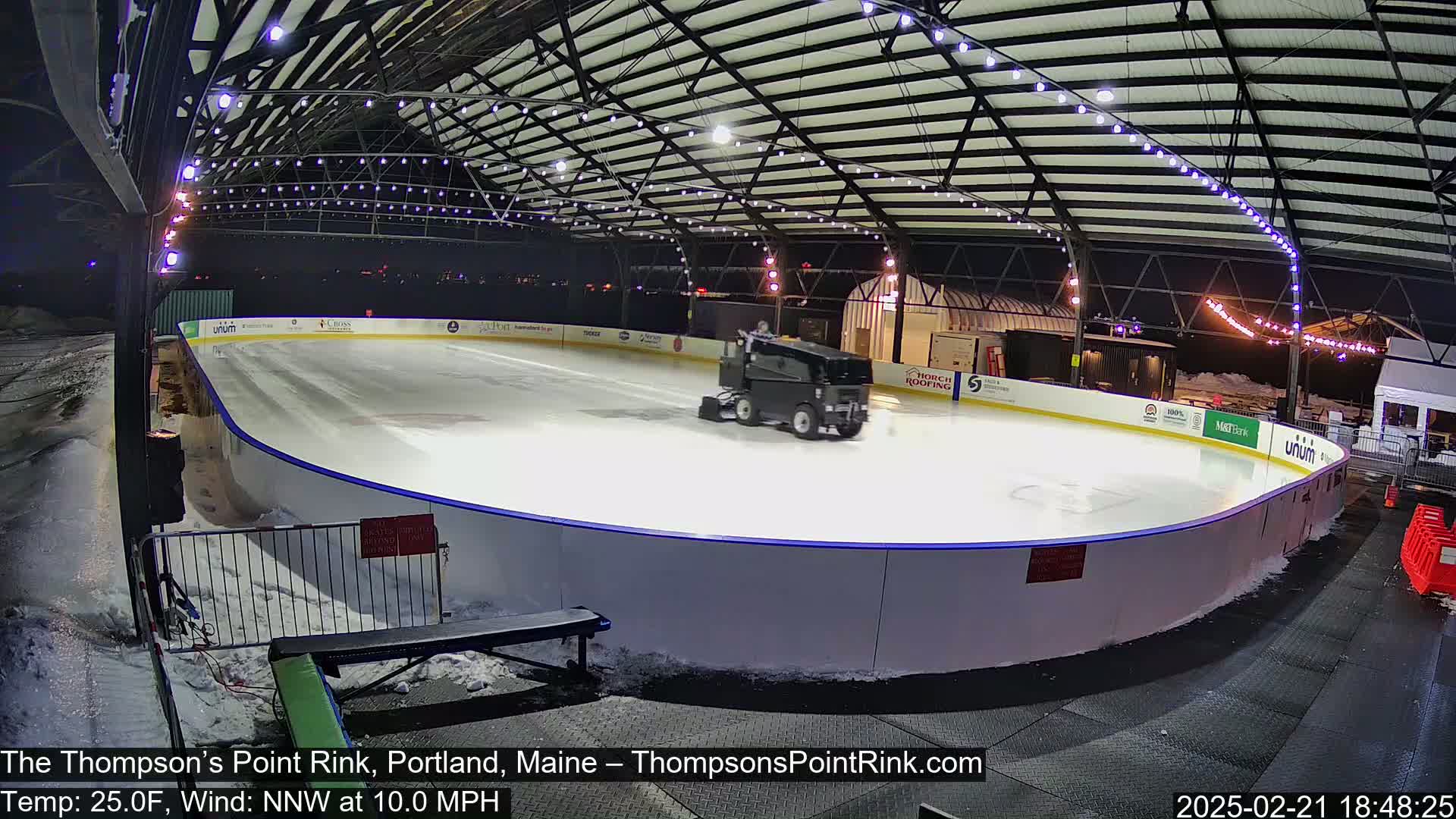 A Zamboni smooths an outdoor ice rink at night under a metal structure lit with string lights, with a temperature of 25 degrees Fahrenheit and a northwest wind of 10 mph.