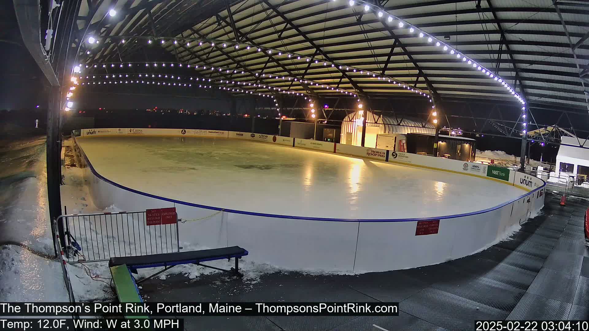An outdoor ice rink, illuminated at night, is surrounded by snow and a partially enclosed structure with string lights.