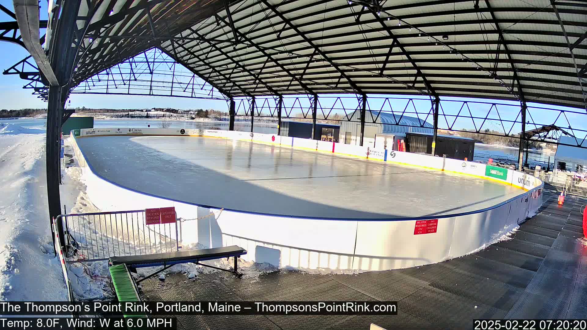 An outdoor ice rink, under a metal roof and partially surrounded by snow, on a sunny day with a temperature of 8°F and a 6 mph west wind.