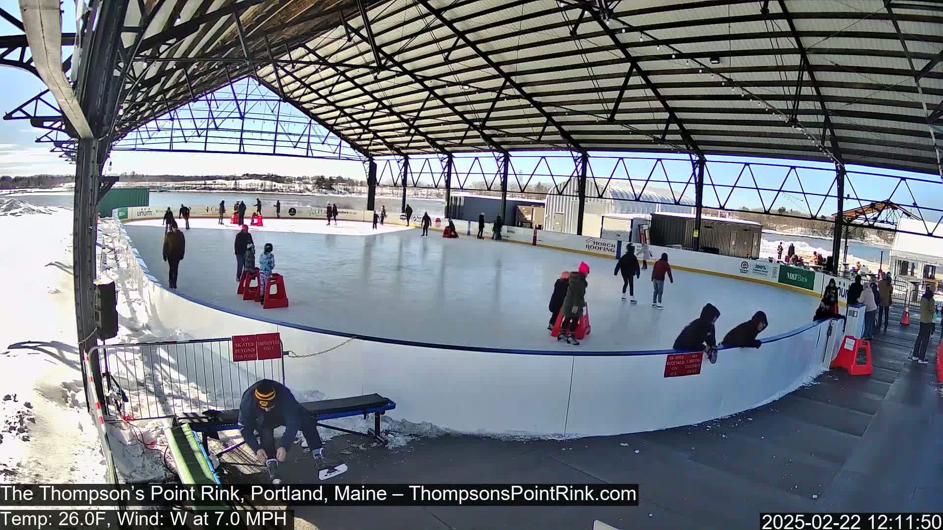 A partially covered outdoor ice rink with several people ice skating on a sunny day.