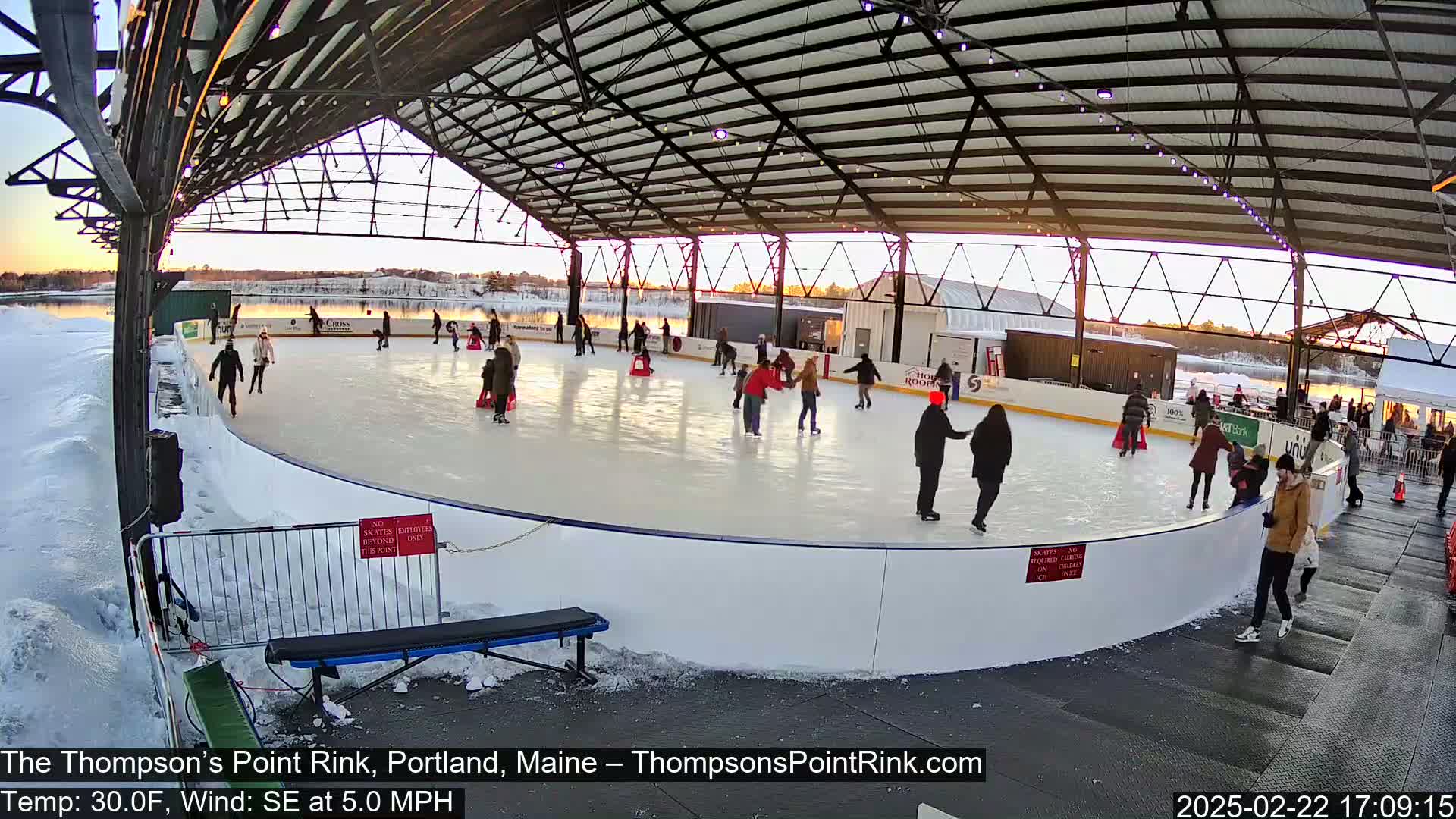 An outdoor ice rink, partially covered by a metal structure, is filled with people ice skating under a partly cloudy sunset sky.