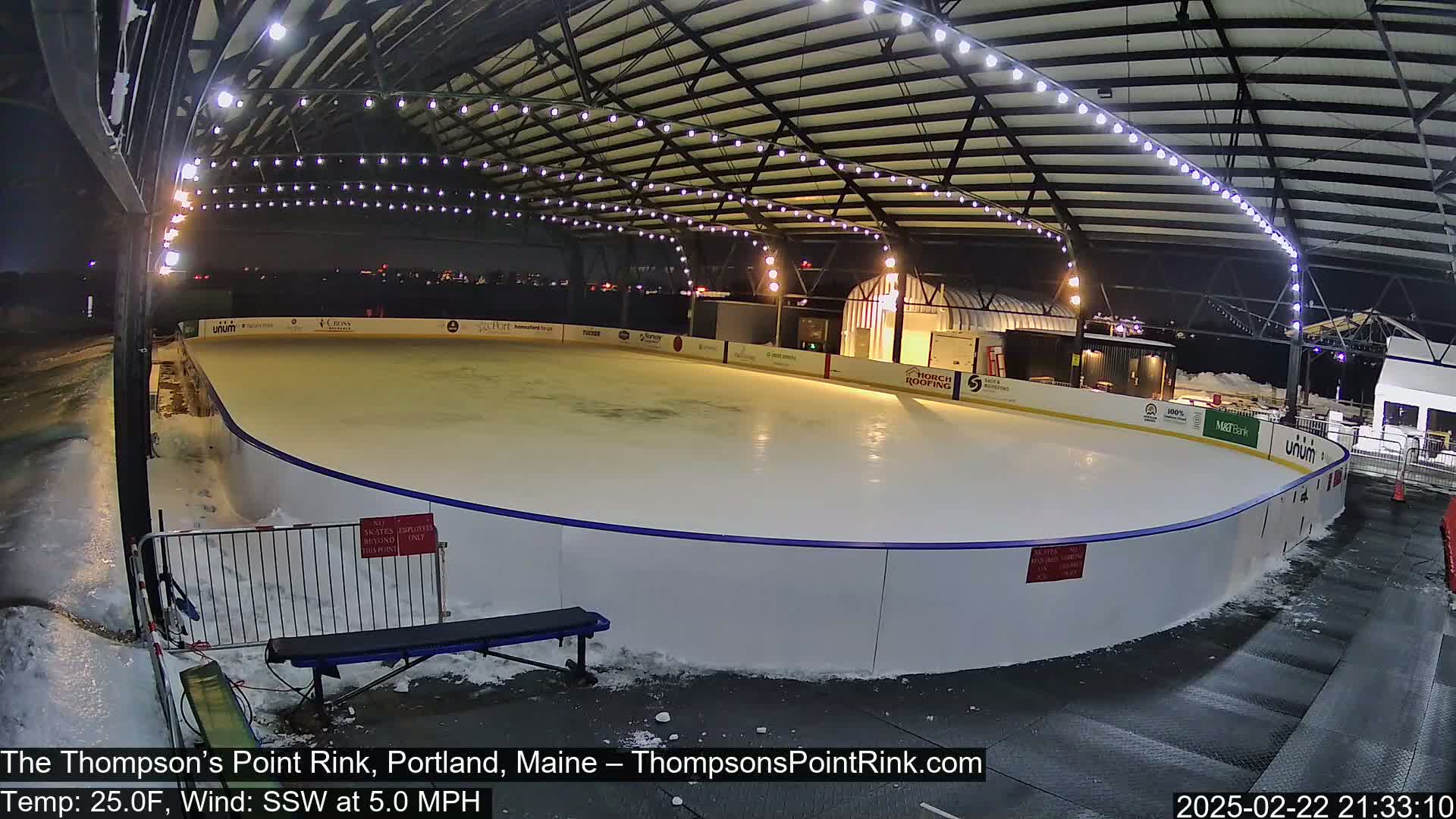 An outdoor ice rink, lit by overhead lights, is surrounded by snow and a metal fence under a clear, nighttime sky.