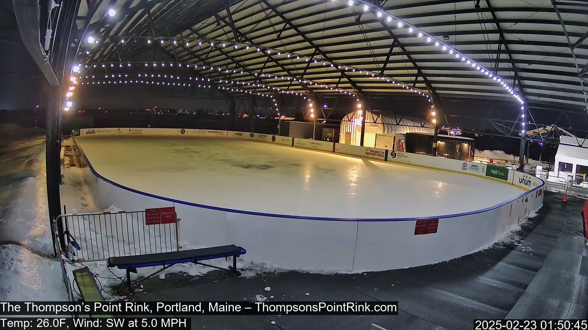 An outdoor ice rink is illuminated at night under a covered structure with snow on the ground and a light southwest wind.