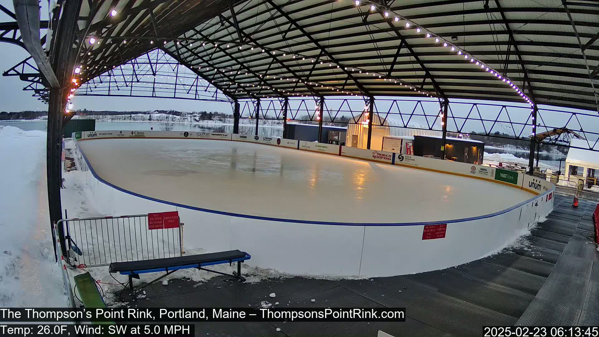 An outdoor ice rink, under a covered structure with string lights, is surrounded by snow in overcast conditions.