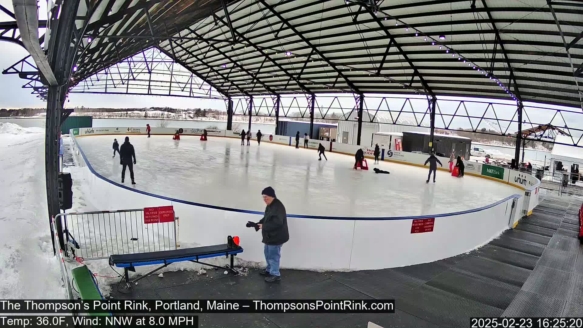 An outdoor ice rink, covered by a metal structure, shows several people ice skating on a partly sunny day with a temperature of 36°F and a northwest wind of 8.0 mph.