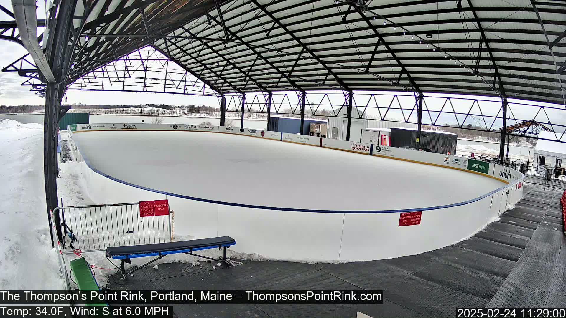 An outdoor ice rink sits under a metal-framed shelter on a partly cloudy day with a light south wind.