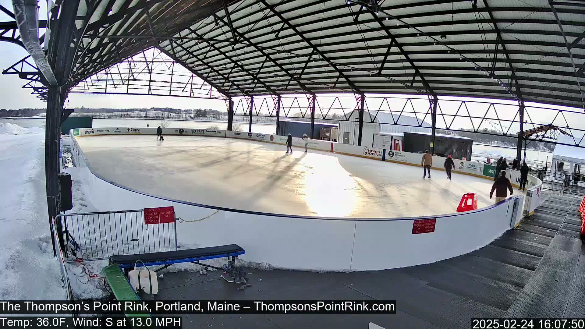 An outdoor ice rink, under a metal structure, with several people ice skating on a sunny day with a temperature of 36°F and a south wind at 13 mph.