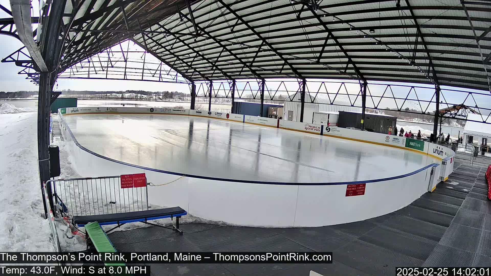 An outdoor ice rink, partially covered by a metal structure, is visible on a sunny day with a south wind.