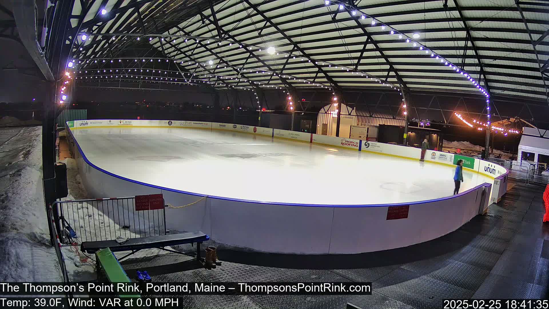 An outdoor ice rink, illuminated at night, is mostly empty except for a few skaters; there is some snow on the ground surrounding the rink.