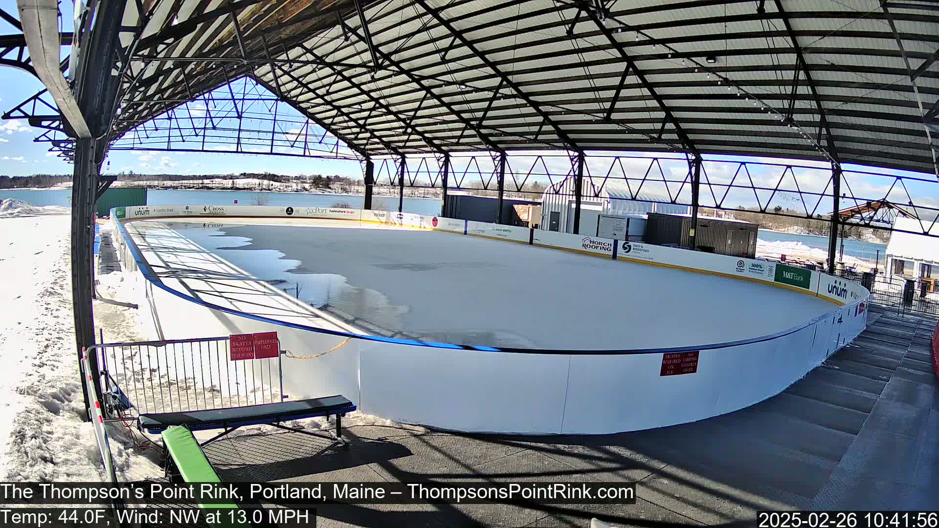 An outdoor ice rink, covered by a metal structure, is partially covered in snow and ice under a sunny sky.