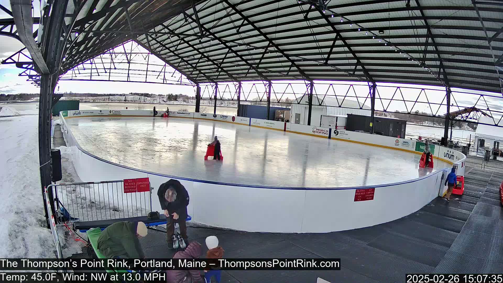 An outdoor ice rink, partially covered by a metal structure, is in use on a partly sunny day with a northwest wind of 13 mph and a temperature of 45°F.