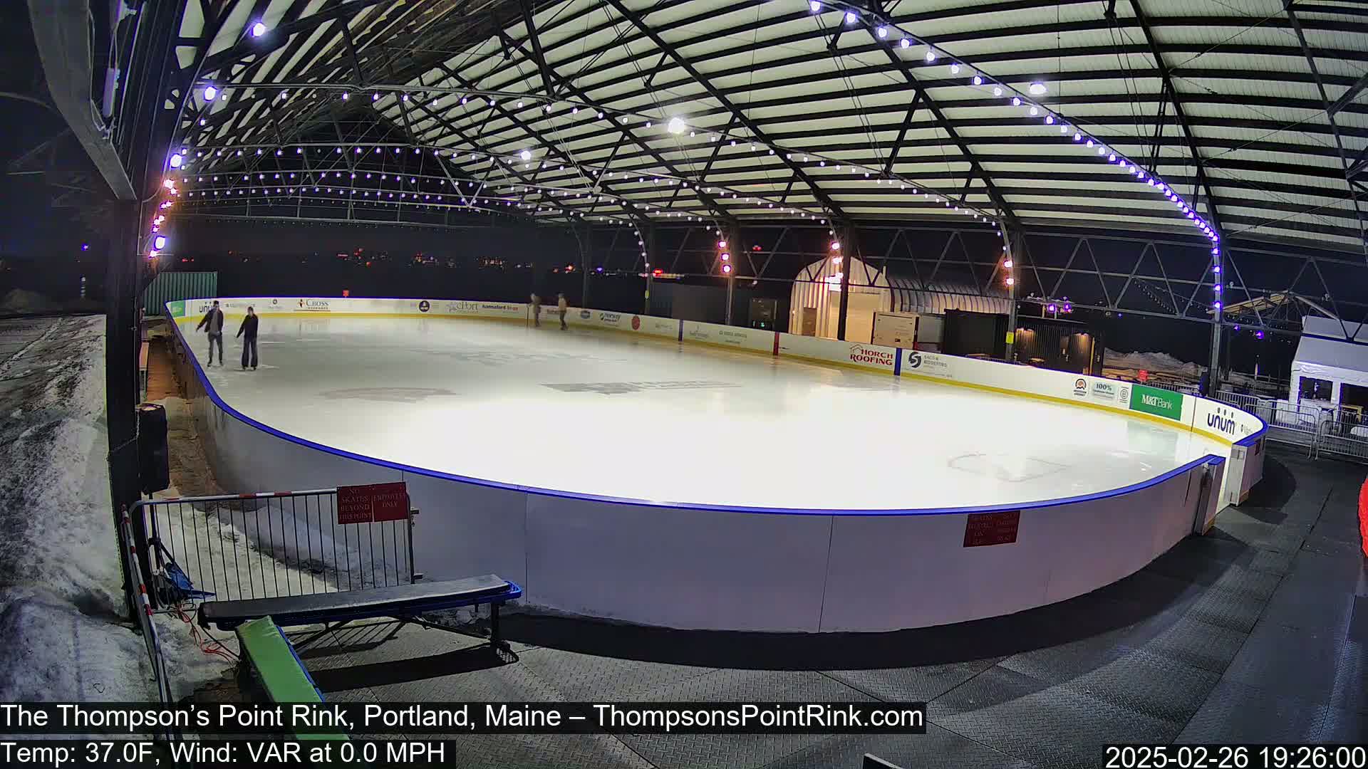 An outdoor ice skating rink, illuminated at night, shows a few people ice skating under a metal structure with string lights; there is snow on the ground surrounding the rink.