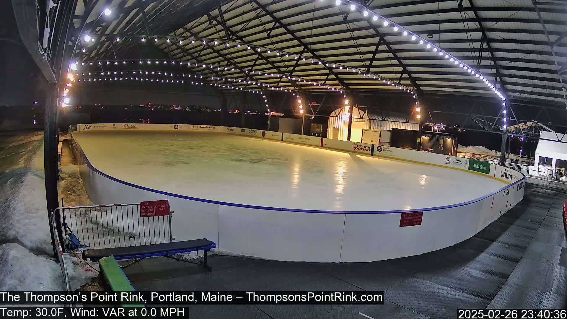 An outdoor ice rink is lit at night under a metal structure, with snow on the ground around it.