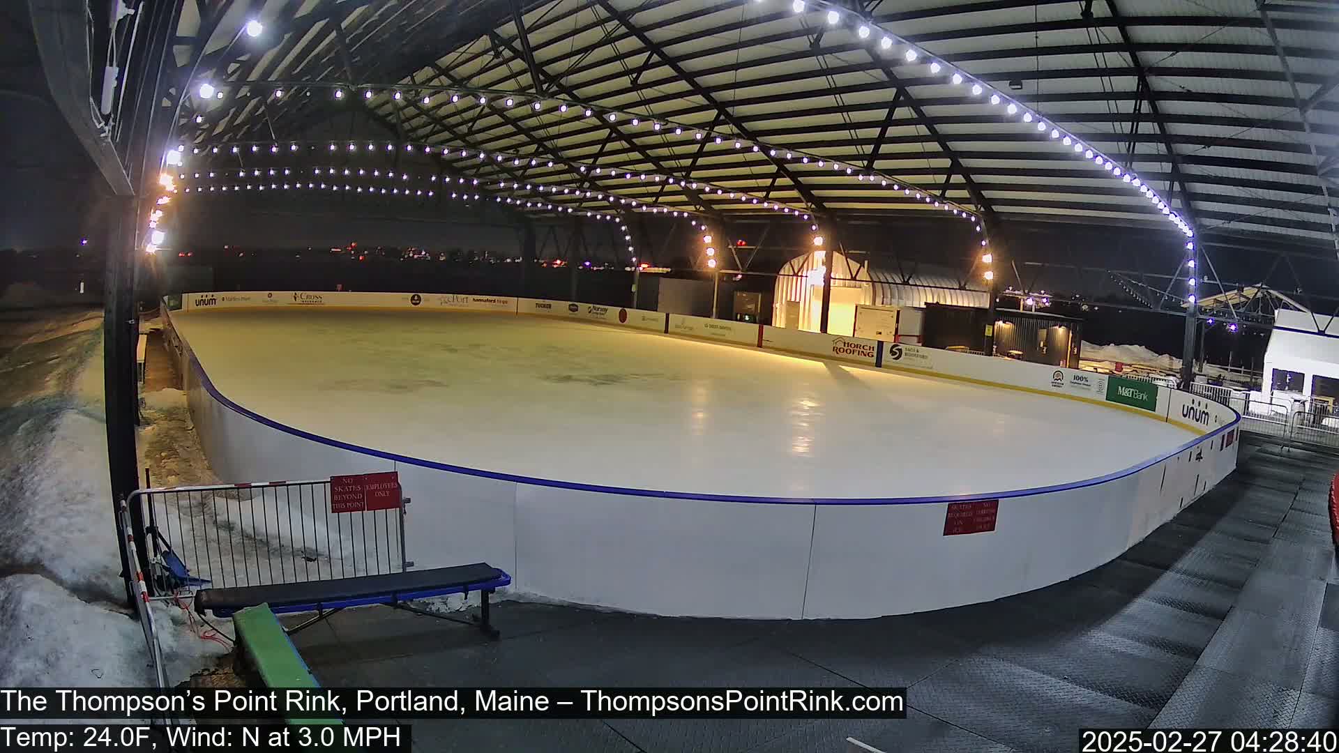 An outdoor ice rink is illuminated at night under a metal roof, with a light snow cover visible outside the rink and a light north wind.
