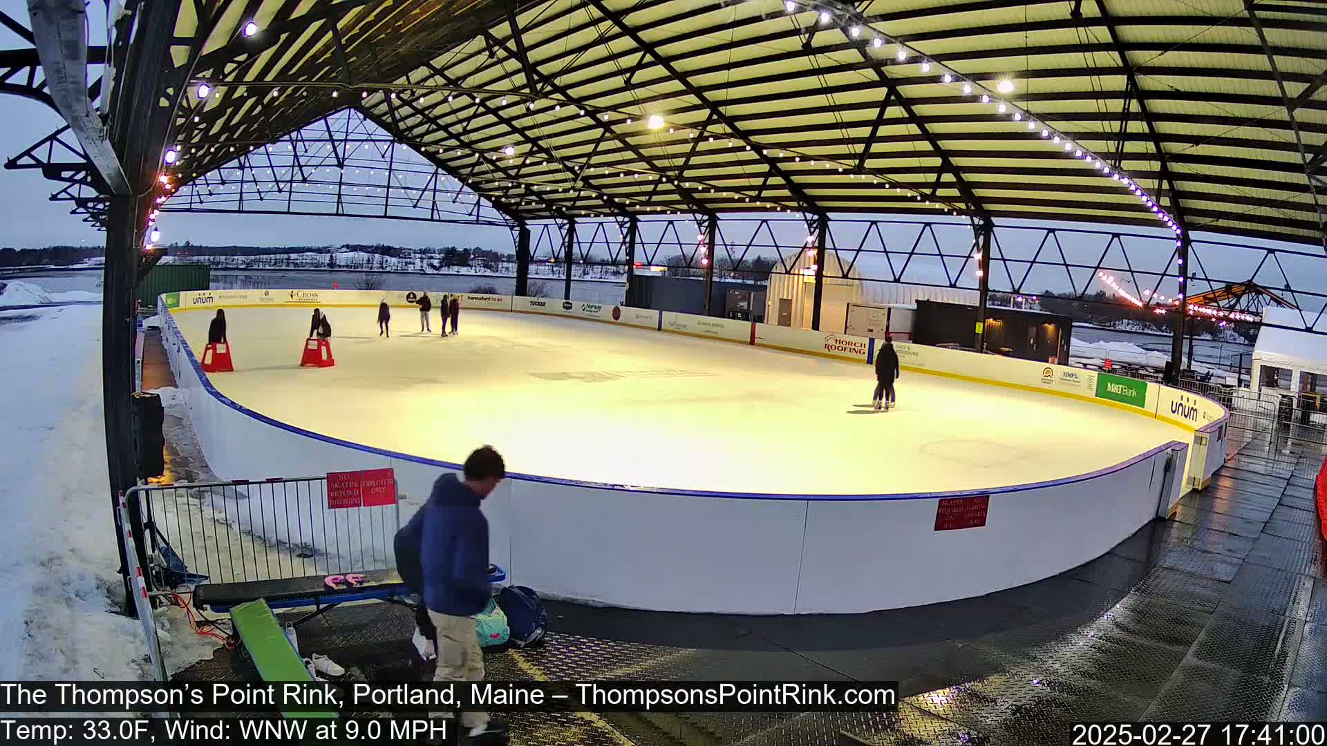 An outdoor ice rink under a metal structure is lit at dusk, with several people ice skating and others standing near the rink's edge.