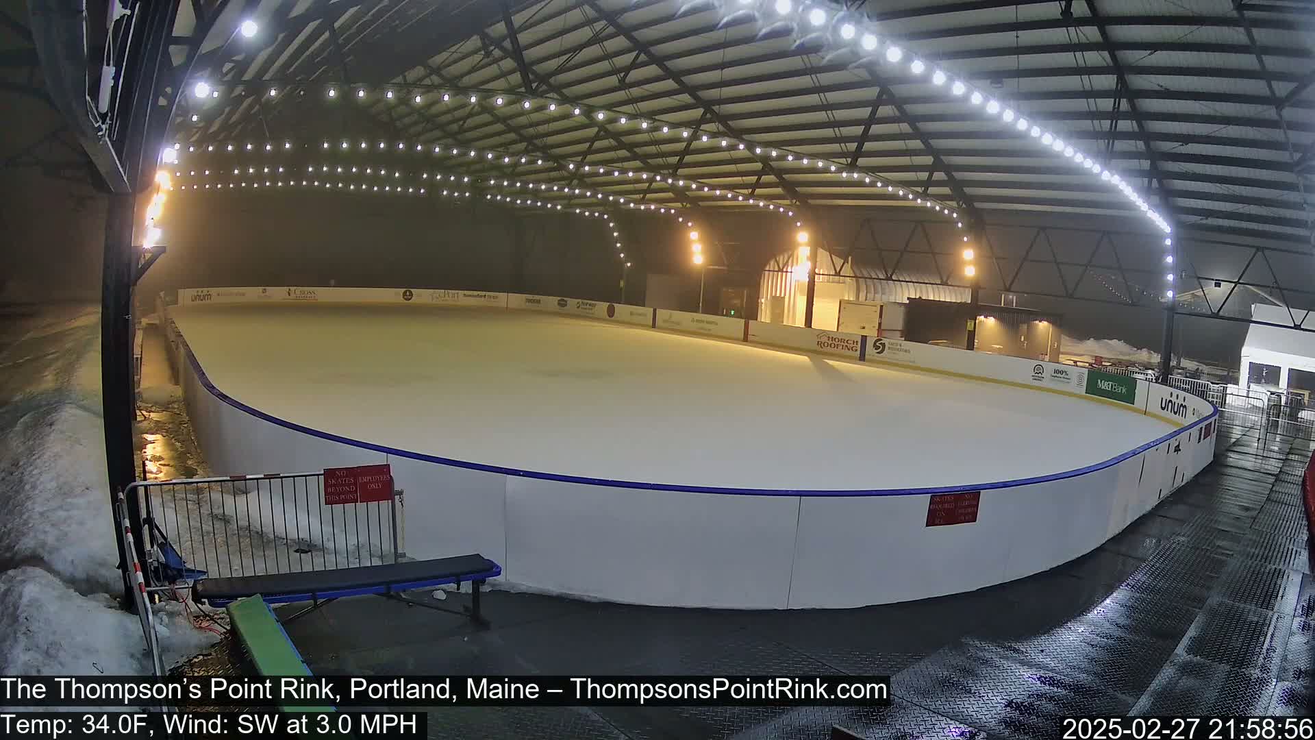 A brightly lit outdoor ice rink is partially covered in snow with a light mist in the air.