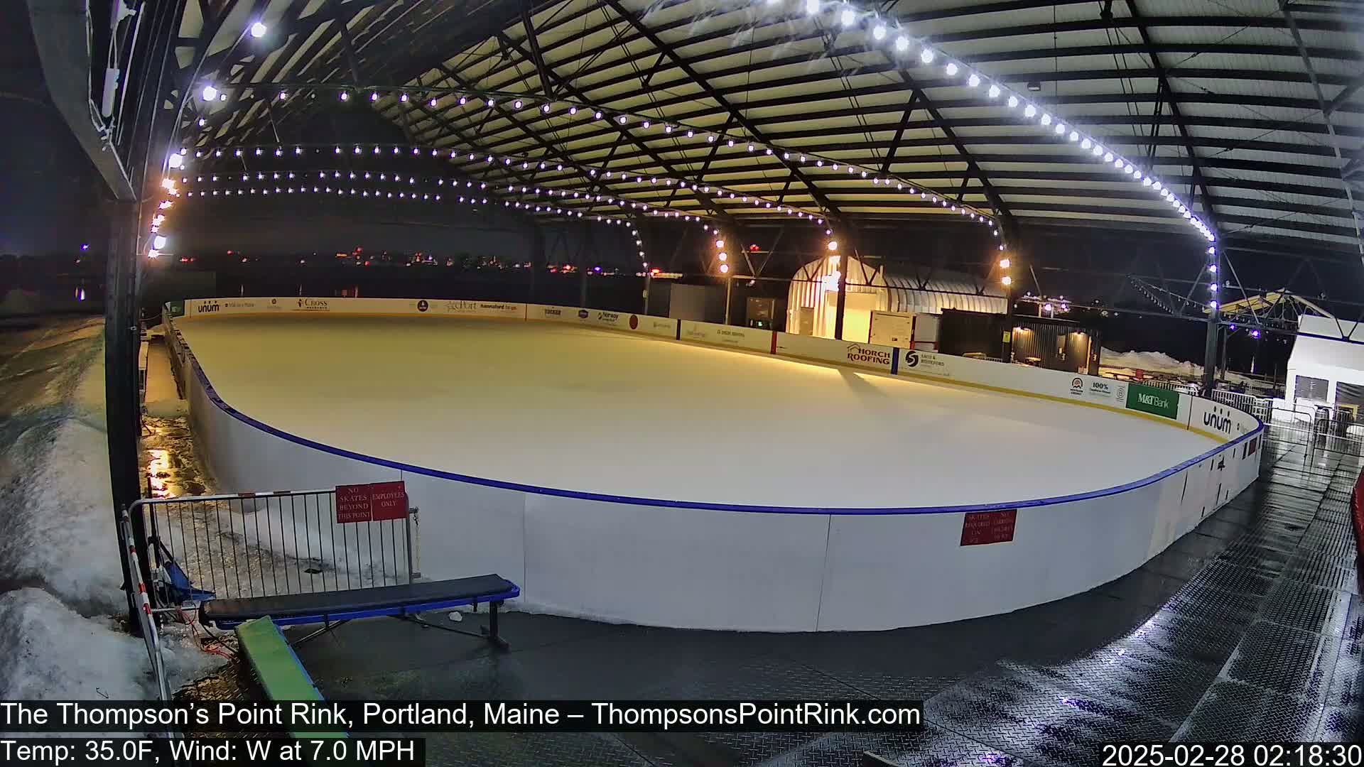 An outdoor ice rink is illuminated at night under a metal structure during a cold, windy evening with some snow and ice visible around the rink.