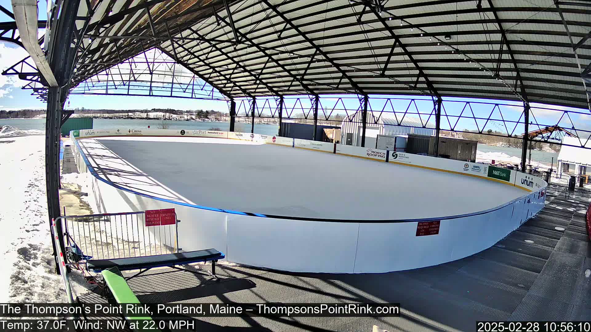 A snow-covered outdoor ice rink sits under a metal-framed structure on a sunny day with a northwest wind.
