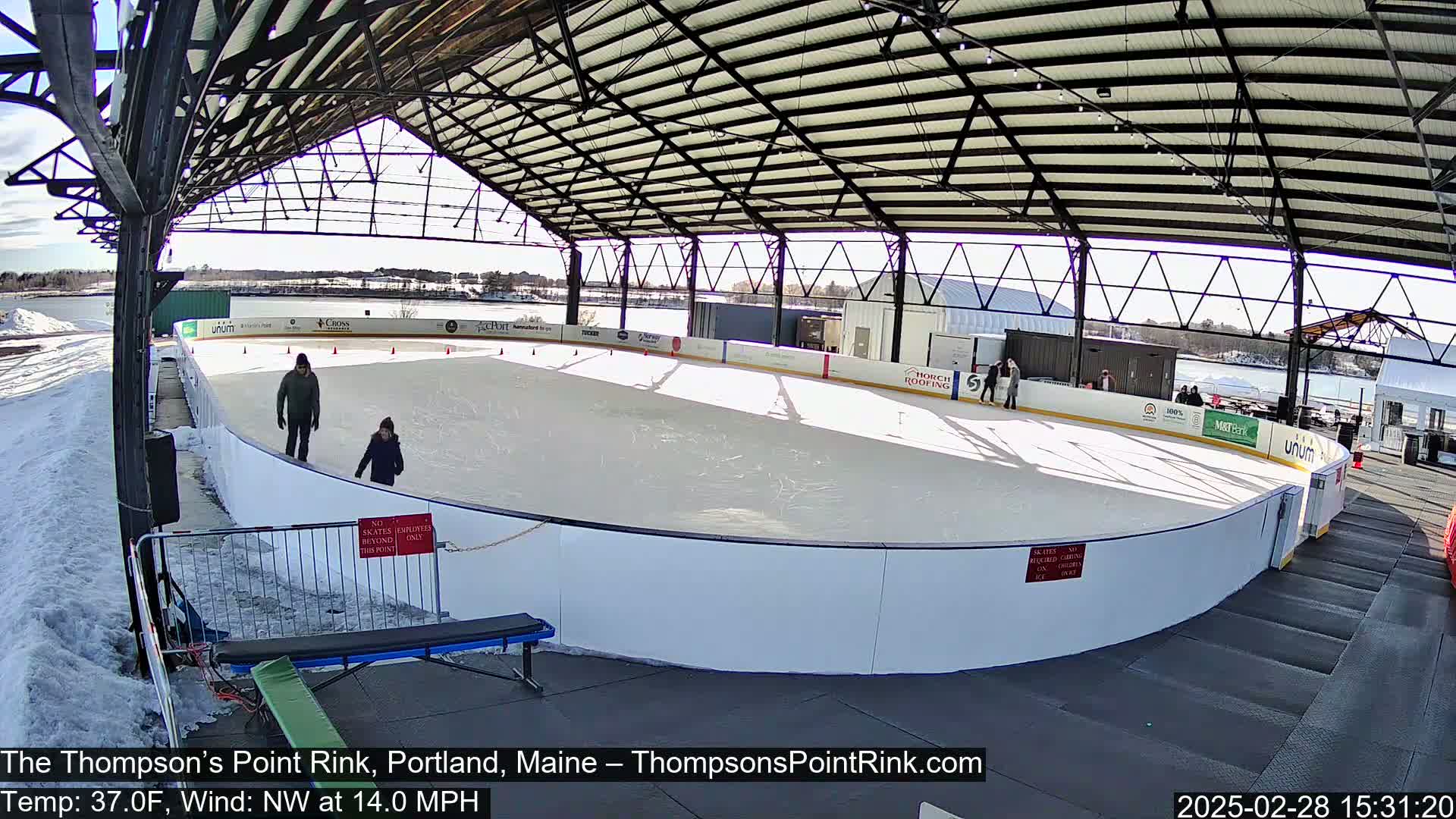 Under a sunny sky, two people skate on an outdoor ice rink enclosed by a metal structure.