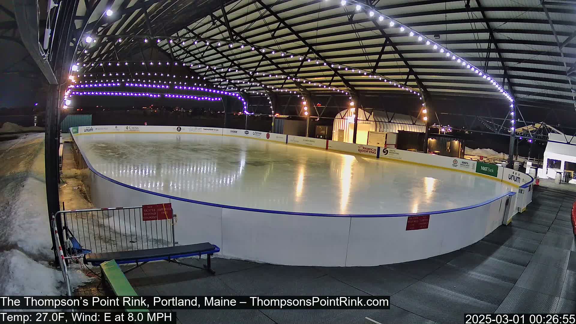 An outdoor ice rink at night, illuminated by string lights, with a slight easterly wind and a temperature of 27 degrees Fahrenheit.