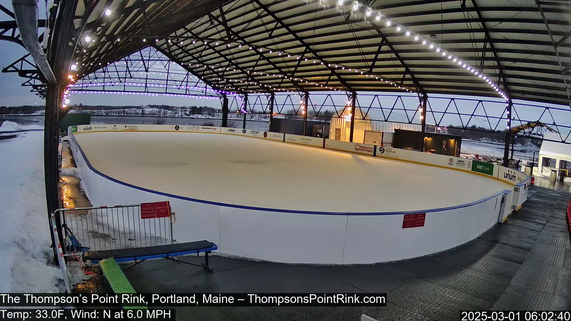 An outdoor ice rink, under a metal structure with string lights, is partially covered in snow, with a mostly clear sky and a light wind.