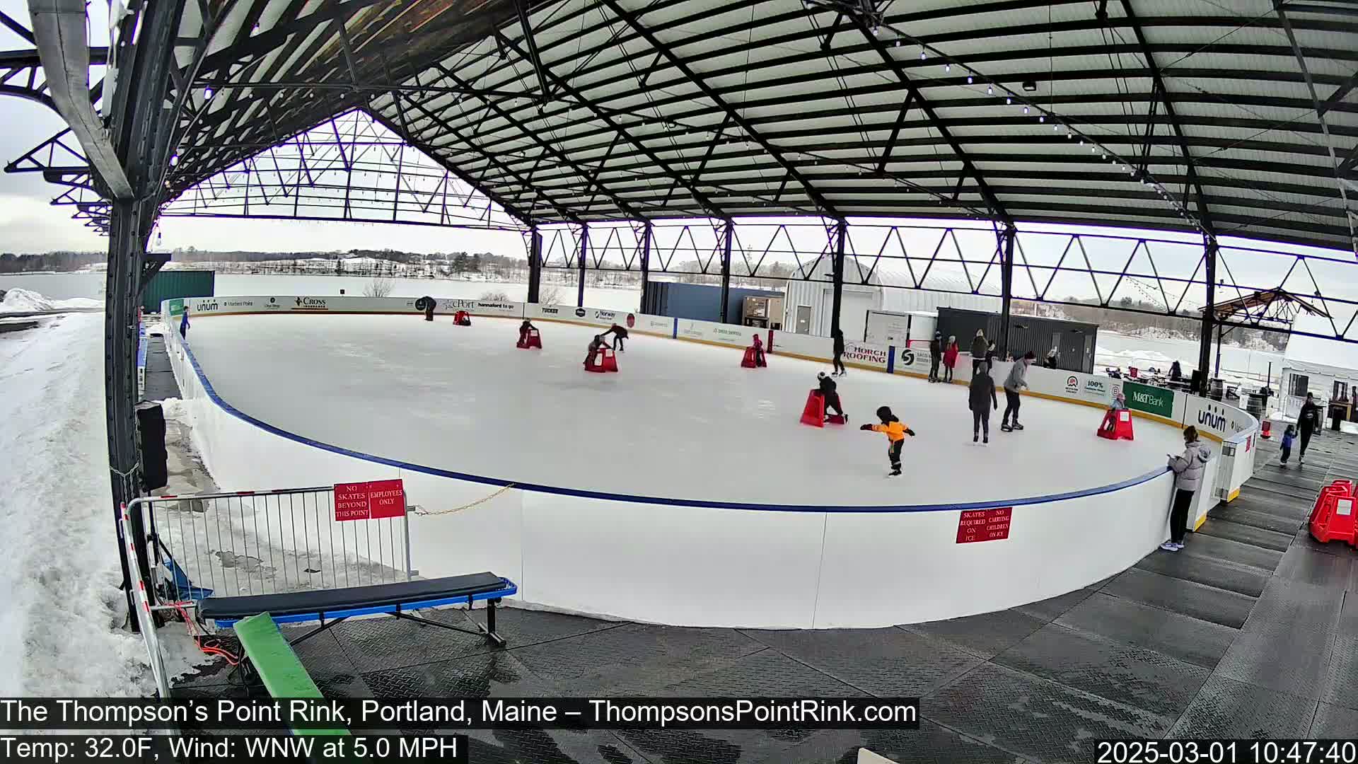 An outdoor ice rink, partially covered by a metal structure, is filled with people ice skating on a cold, partly cloudy day.