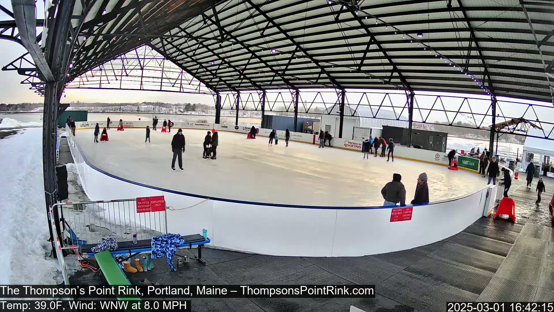An outdoor ice skating rink under a metal roof structure on a partly cloudy day with a temperature of 39 degrees Fahrenheit and an 8 mph wind.