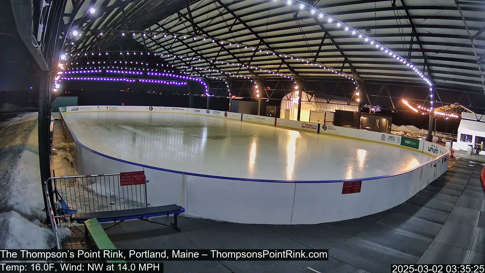 An outdoor ice rink, illuminated at night under a metal structure with string lights, is partially covered in snow with a northwest wind at 14 mph and a temperature of 16°F.