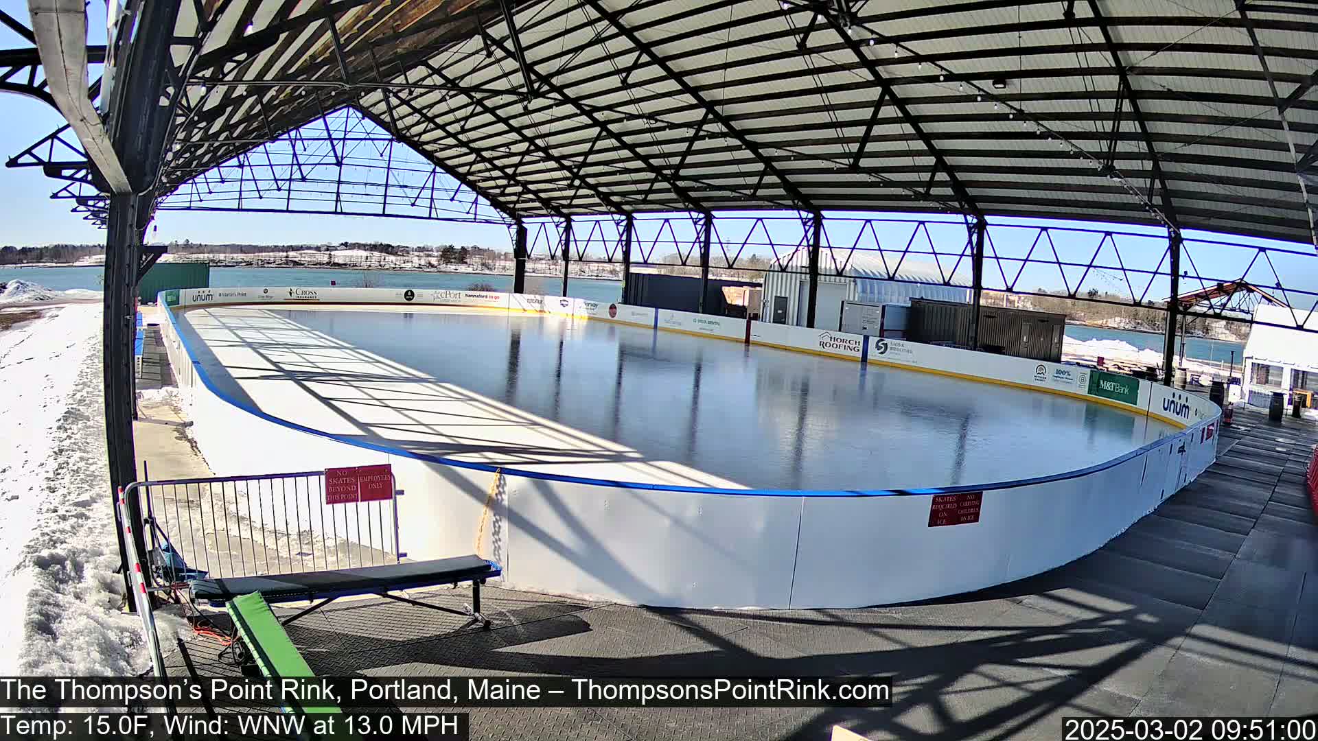 A sunny outdoor ice rink sits under a metal-framed roof with a body of water visible in the background.