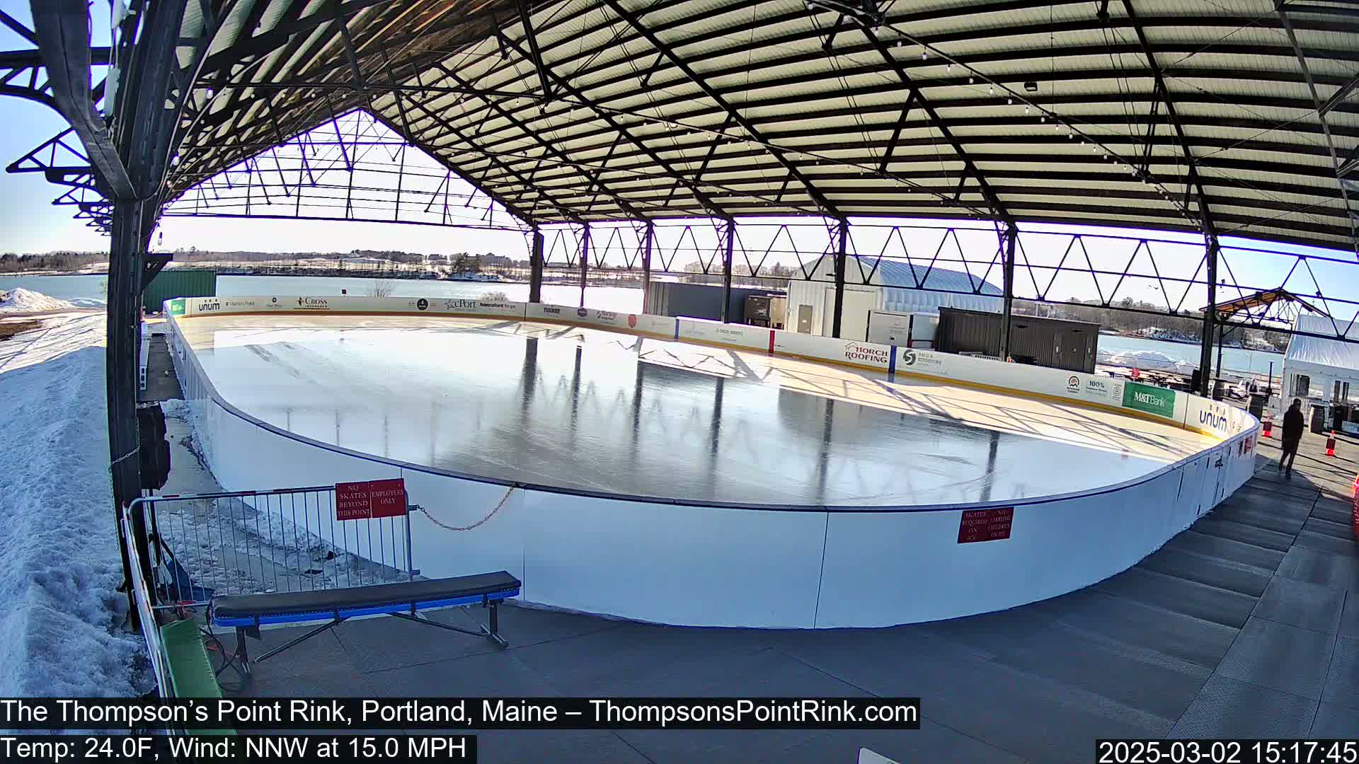An outdoor ice rink, enclosed under a metal framework, is visible on a sunny day with a northwest wind of 15 mph and a temperature of 24 degrees Fahrenheit.