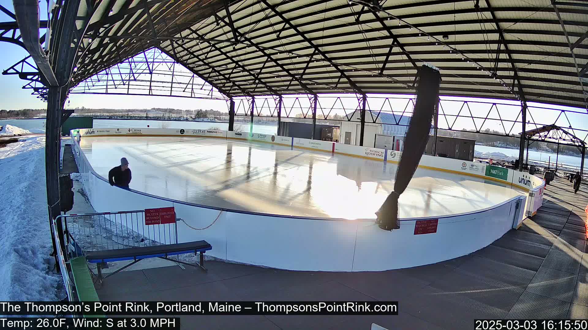 An outdoor ice rink, covered by a metal structure, is partially surrounded by snow, on a sunny day with a south wind of 3 mph and a temperature of 26 degrees Fahrenheit.