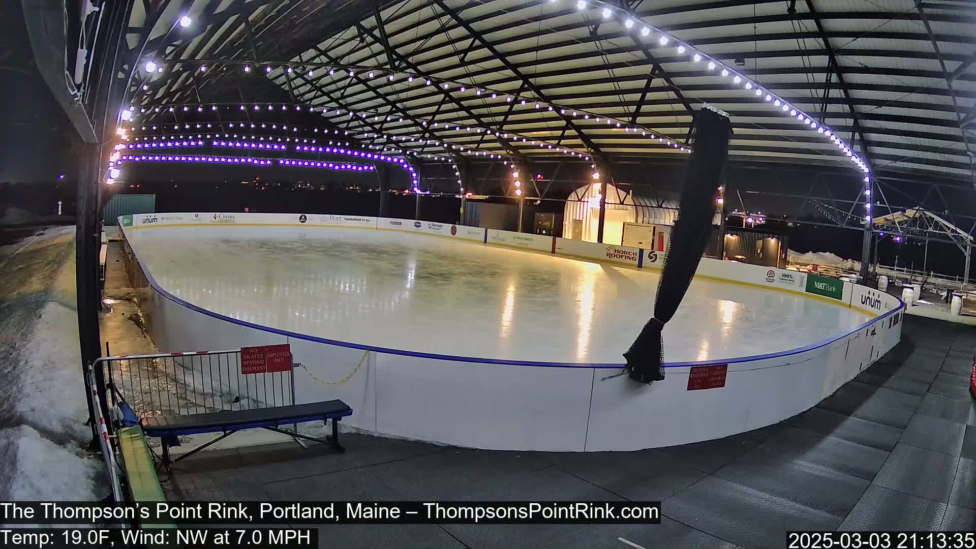 An outdoor ice rink, lit at night, is mostly empty with some minor snow and ice buildup around the rink perimeter; the temperature is 19°F and the wind is from the northwest at 7 mph.