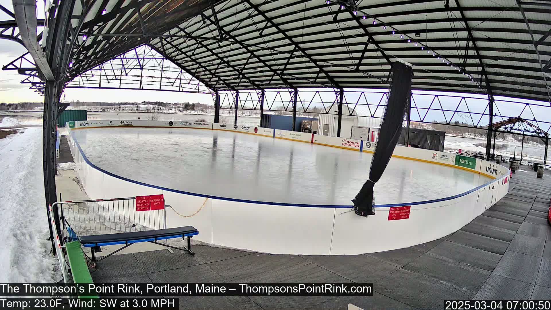 An outdoor ice rink, covered by a metal structure, is visible on a partly cloudy day with a temperature of 23 degrees Fahrenheit and a southwest wind of 3 miles per hour.