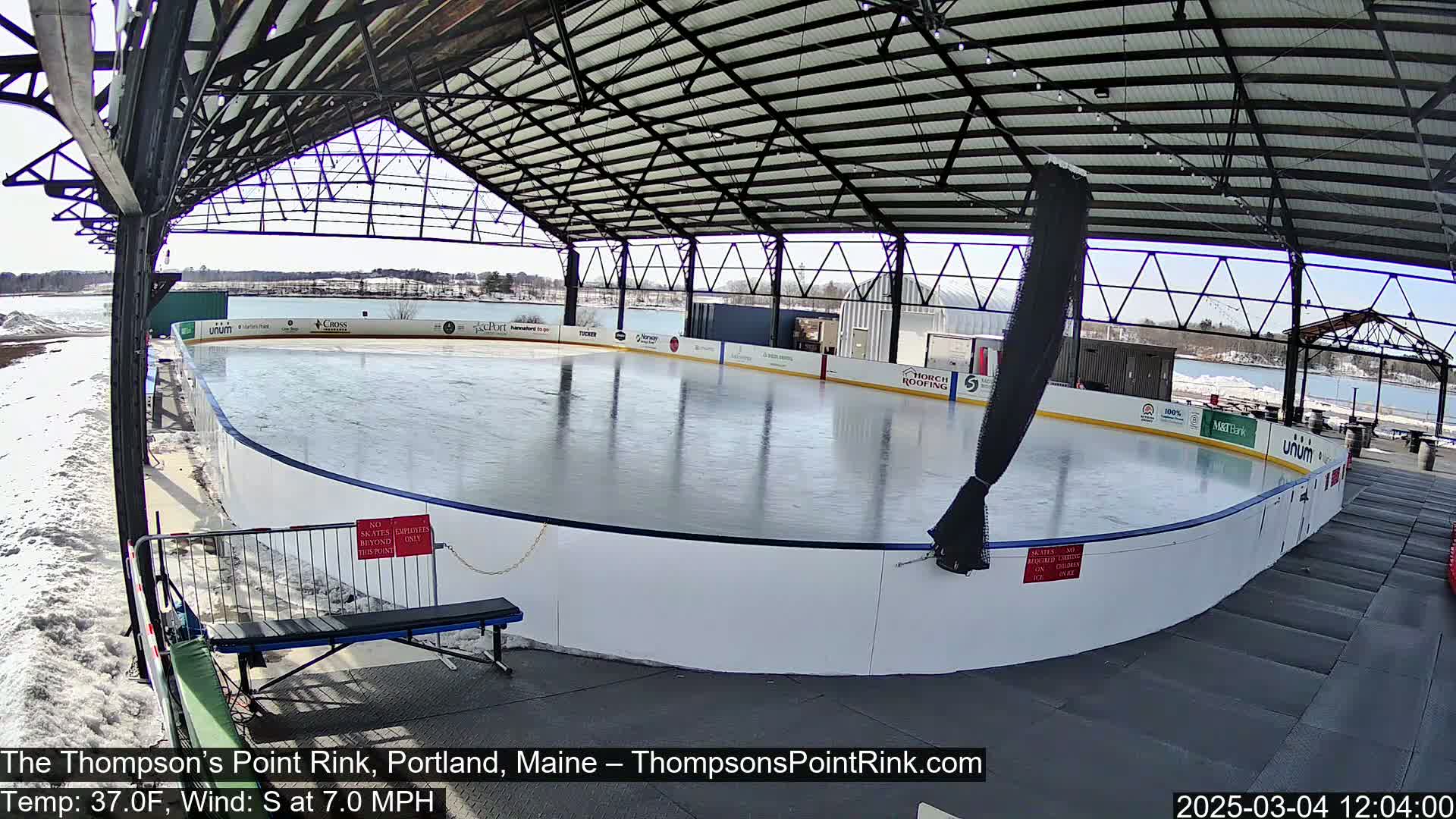 An outdoor ice rink, covered by a metal structure, sits in a snowy area under a sunny sky.