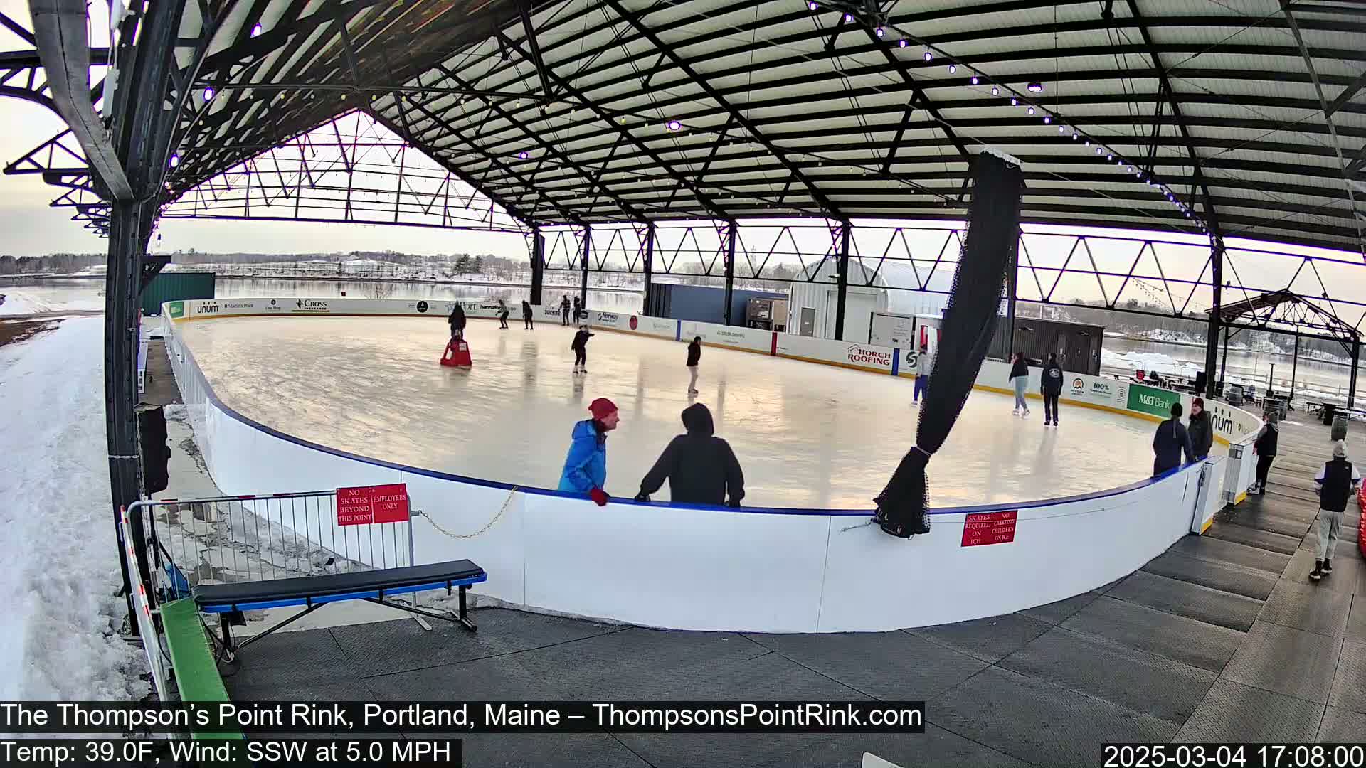 An outdoor ice rink, under a metal structure, with several people ice skating on a partly sunny day, with a temperature of 39°F and a southwest wind of 5 mph.
