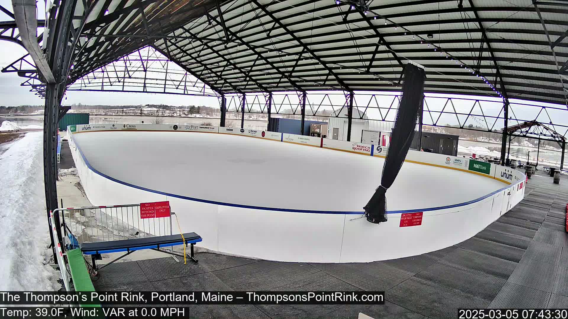 An outdoor ice rink, under a metal structure, is partially covered in snow, with a partly cloudy sky above.