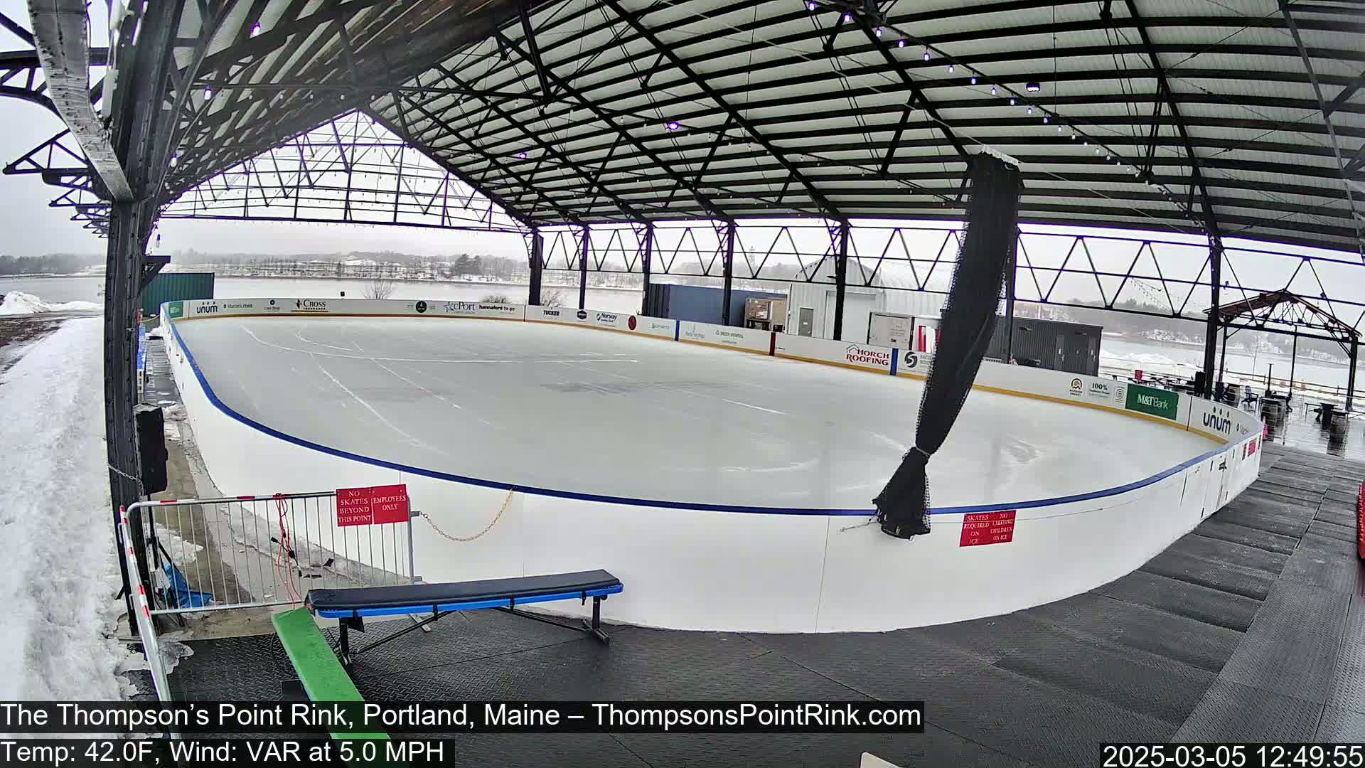 An outdoor ice rink under a metal-framed structure on a snowy, overcast day.