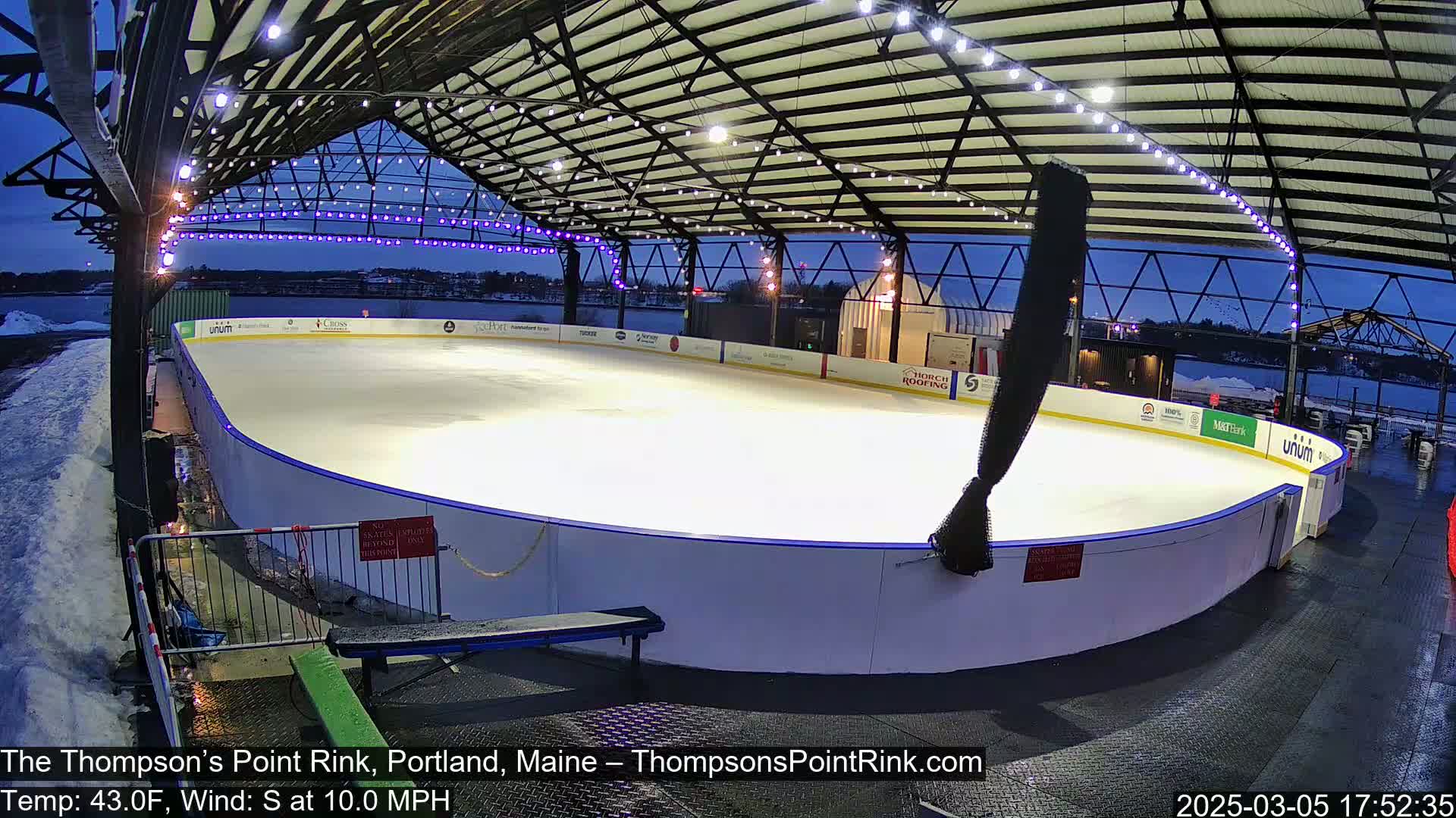 An outdoor ice rink, under a metal structure with string lights, is partially covered in snow, at dusk under a twilight sky.