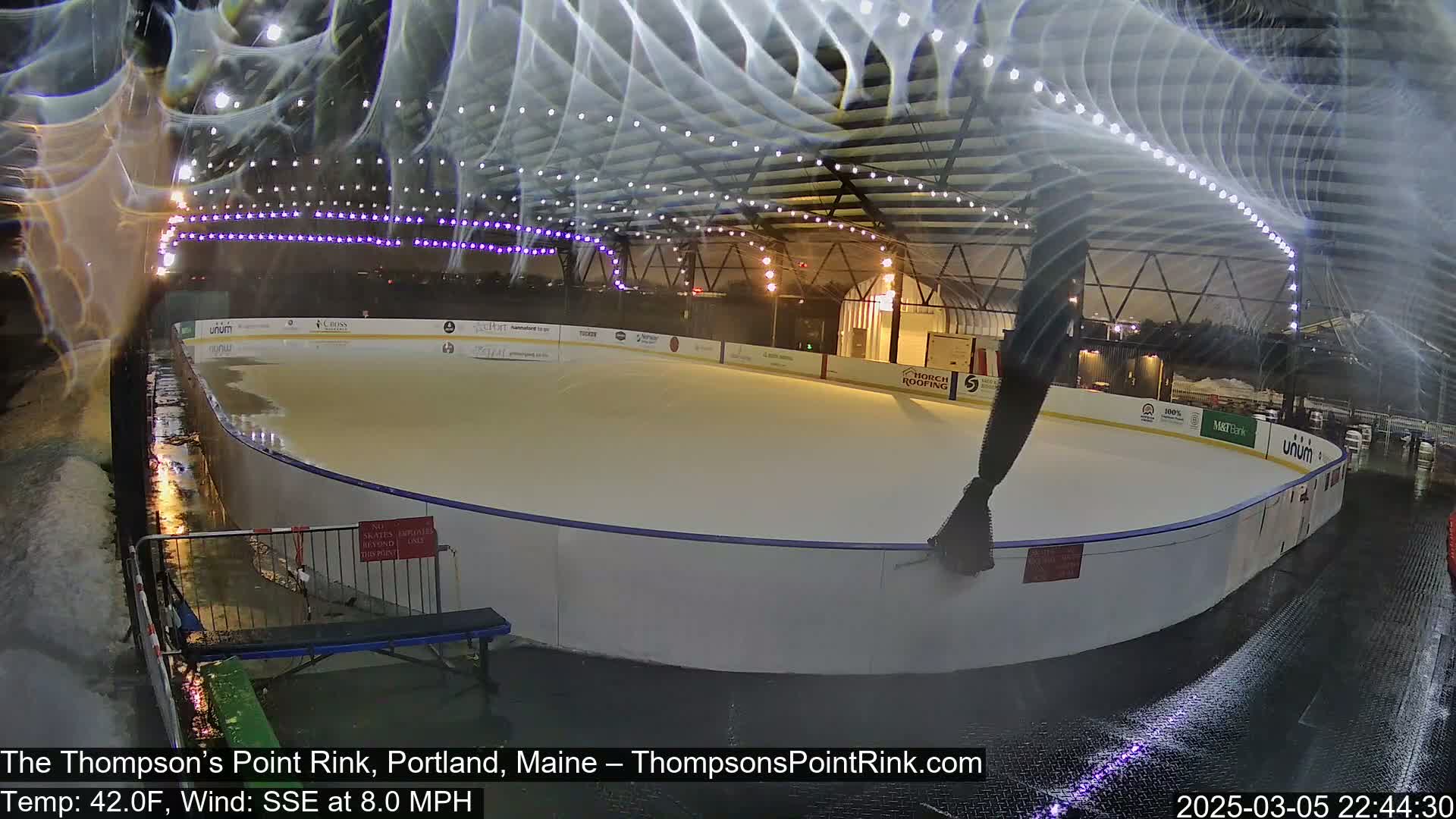 An outdoor ice rink is wet and surrounded by puddles under a dark sky with string lights overhead, in windy conditions.