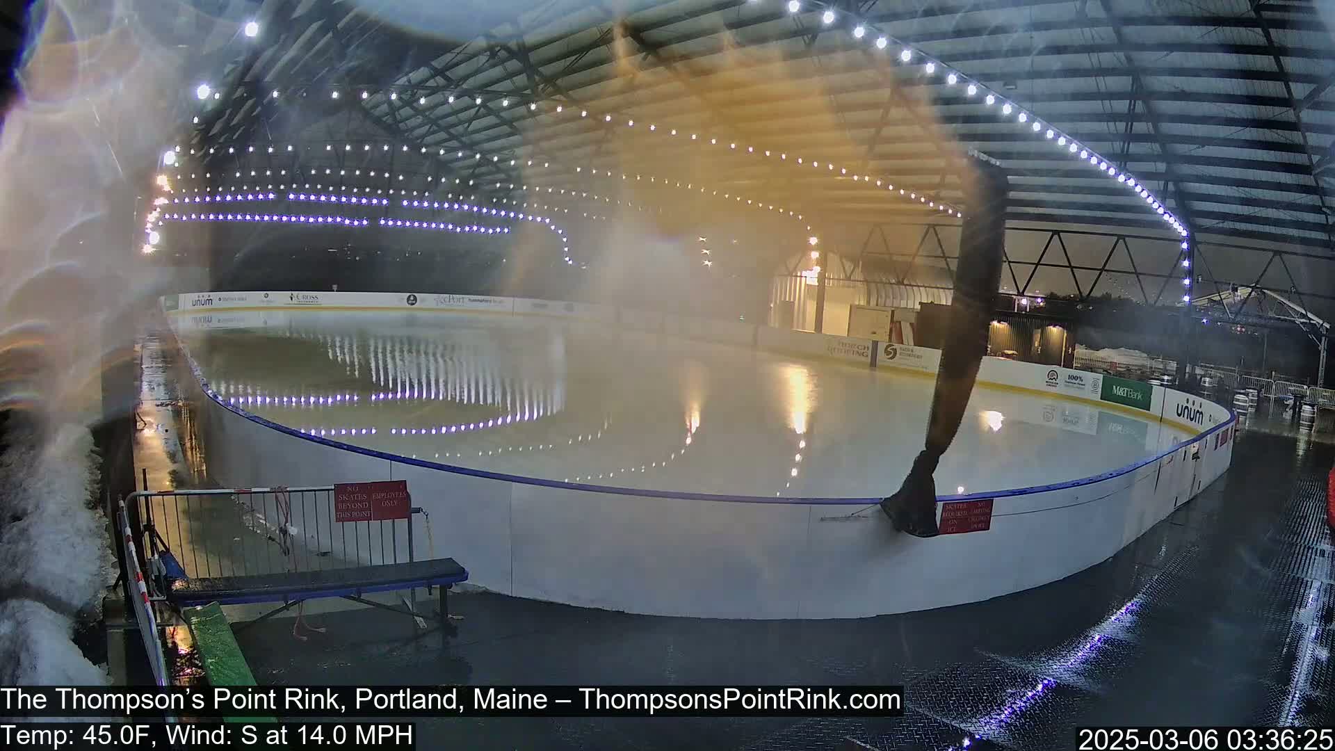 An outdoor ice rink is being sprayed with water in windy, 45°F weather, under a covered structure with overhead lights.